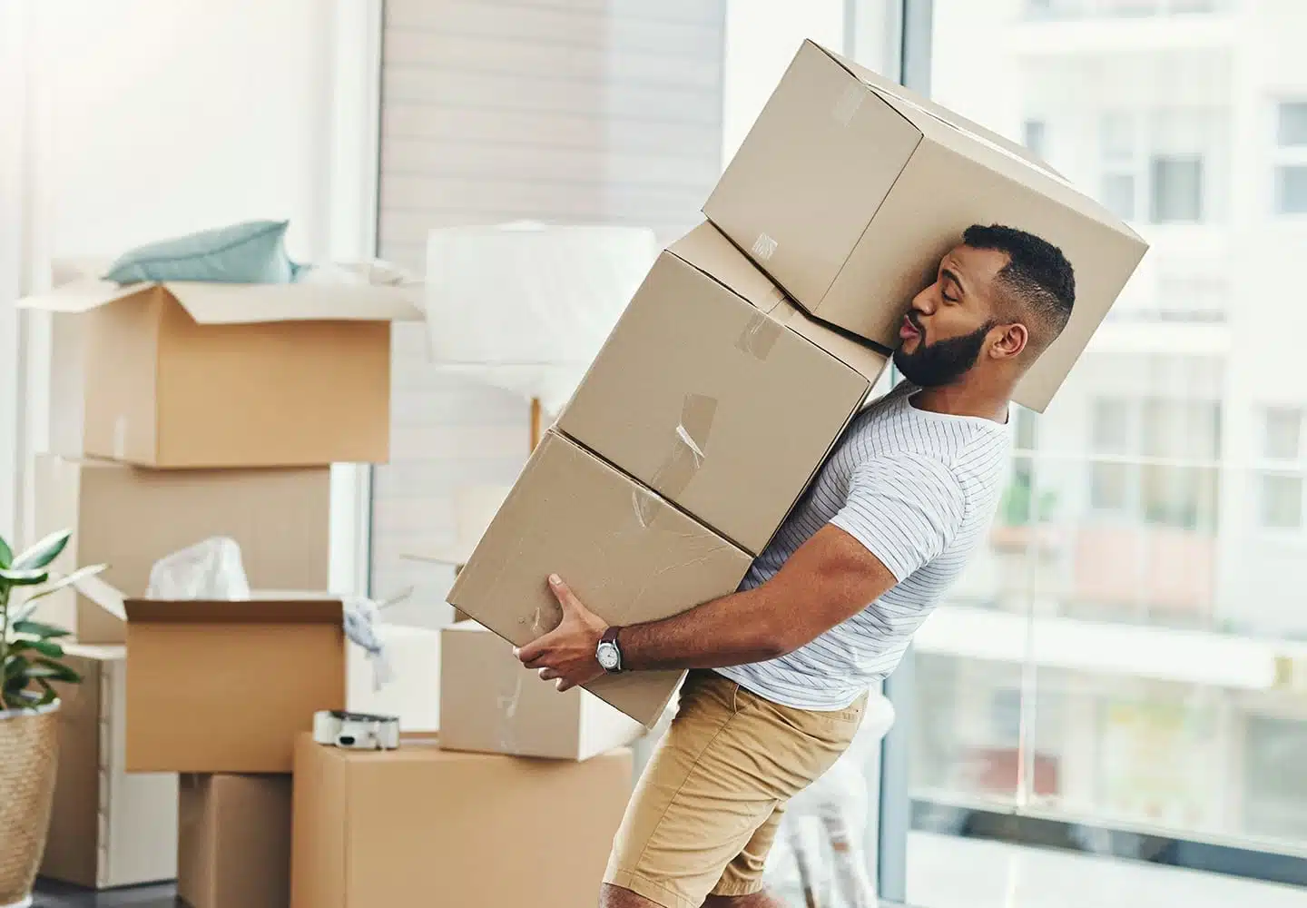 A man carries three cardboard boxes out of his apartment. There are six more stacked being him and a plant is off to the side.