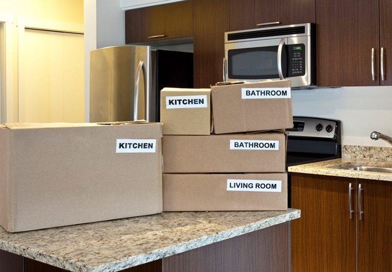 Cardboard boxes sit on a granite counter in a kitchen. They are neatly stacked and labeled "kitchen, kitchen, bathroom, bathroom, living room". 