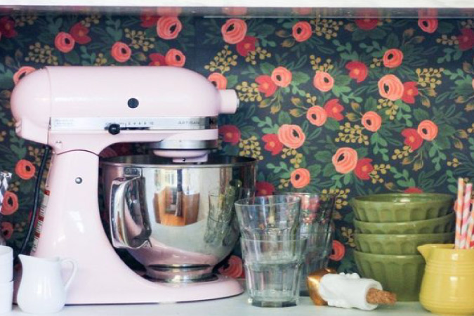 The interior of a cabinet with colorful floral wallpaper on the back wall. There’s a standup mixer and various dishes in the cabinet.