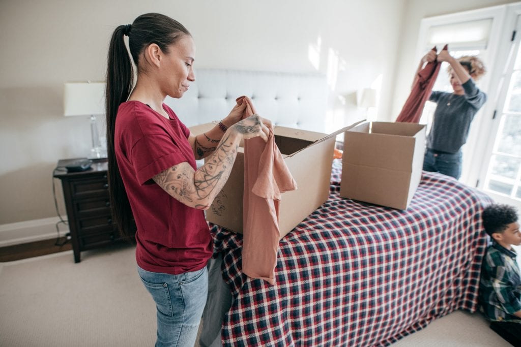 Two young women pack up their apartment to move to their new home. There are boxes that sits on a checkered blanket while their son sits on the floor watching TV. 
