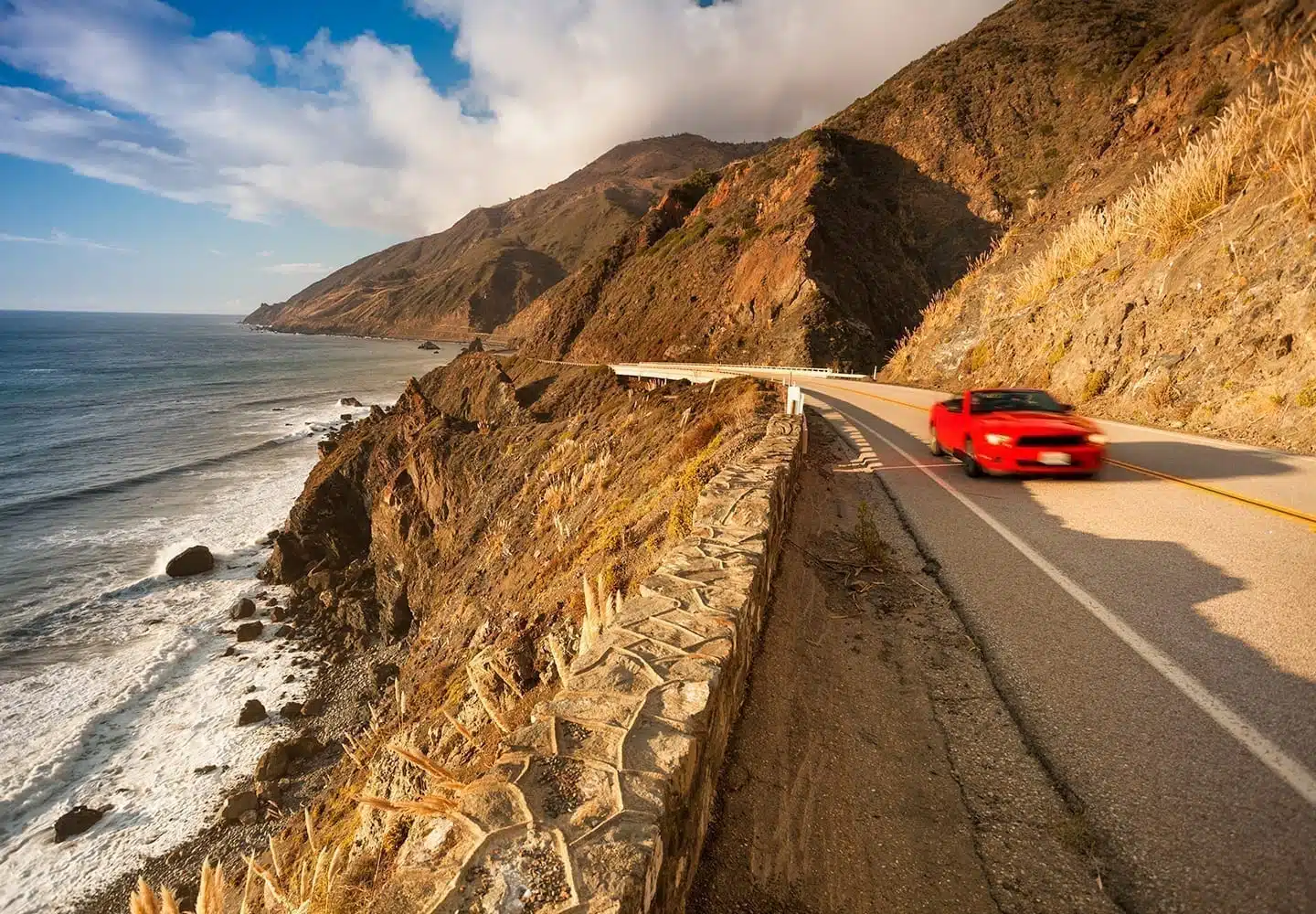 A red Ford Mustang speeds along a mountainside road. There is a cliff with water below it.