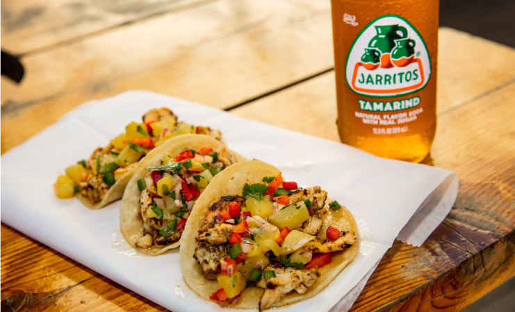 Close up of a wood table with three street tacos and a cold Jarritos Tamarind-flavored soda. The street tacos are resting on top of white paper and are filled with grilled chicken, pico de gallo, and cilantro. 