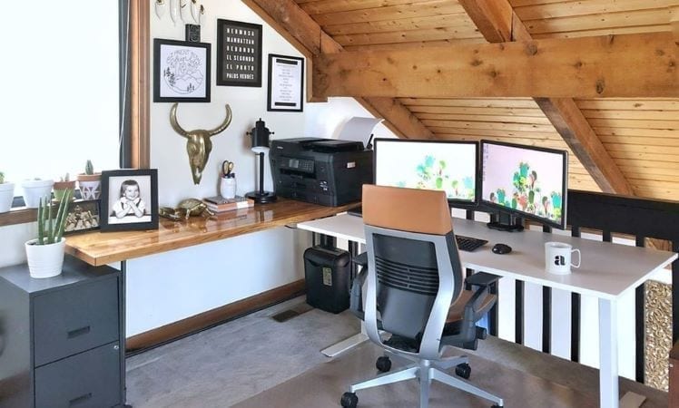 A home office set up in the loft. Two desks have been combined to make a corner desk, art and pictures are hung on the wall, and there are dual computer screens set up for work.