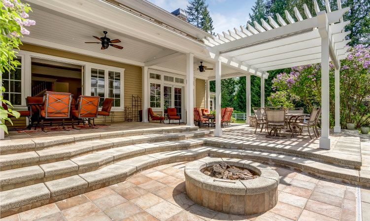 A paved patio in the backyard of a home. It has steps leading down to a fire pit and a pergola casting shade on an outdoor dining set.