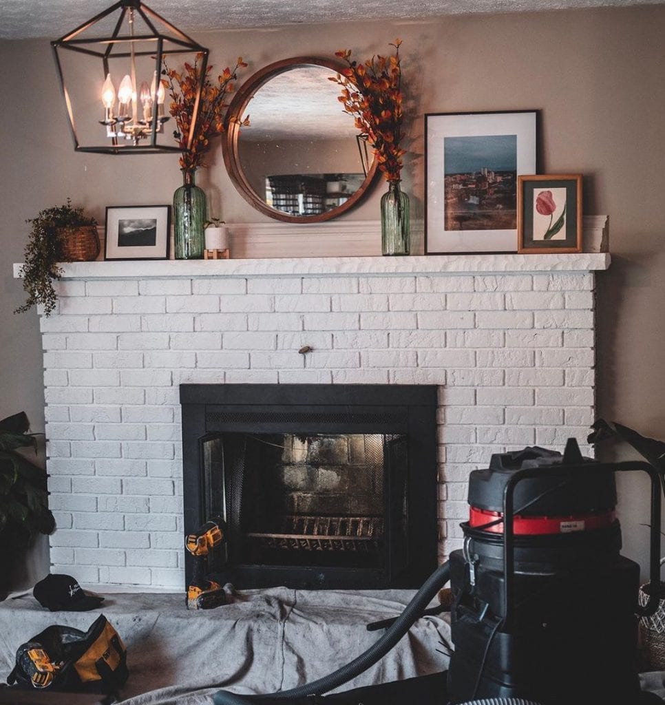 A shop vac, cloth tarp, and tools are placed beside a white brick fireplace as someone prepares to clean it before winter starts.