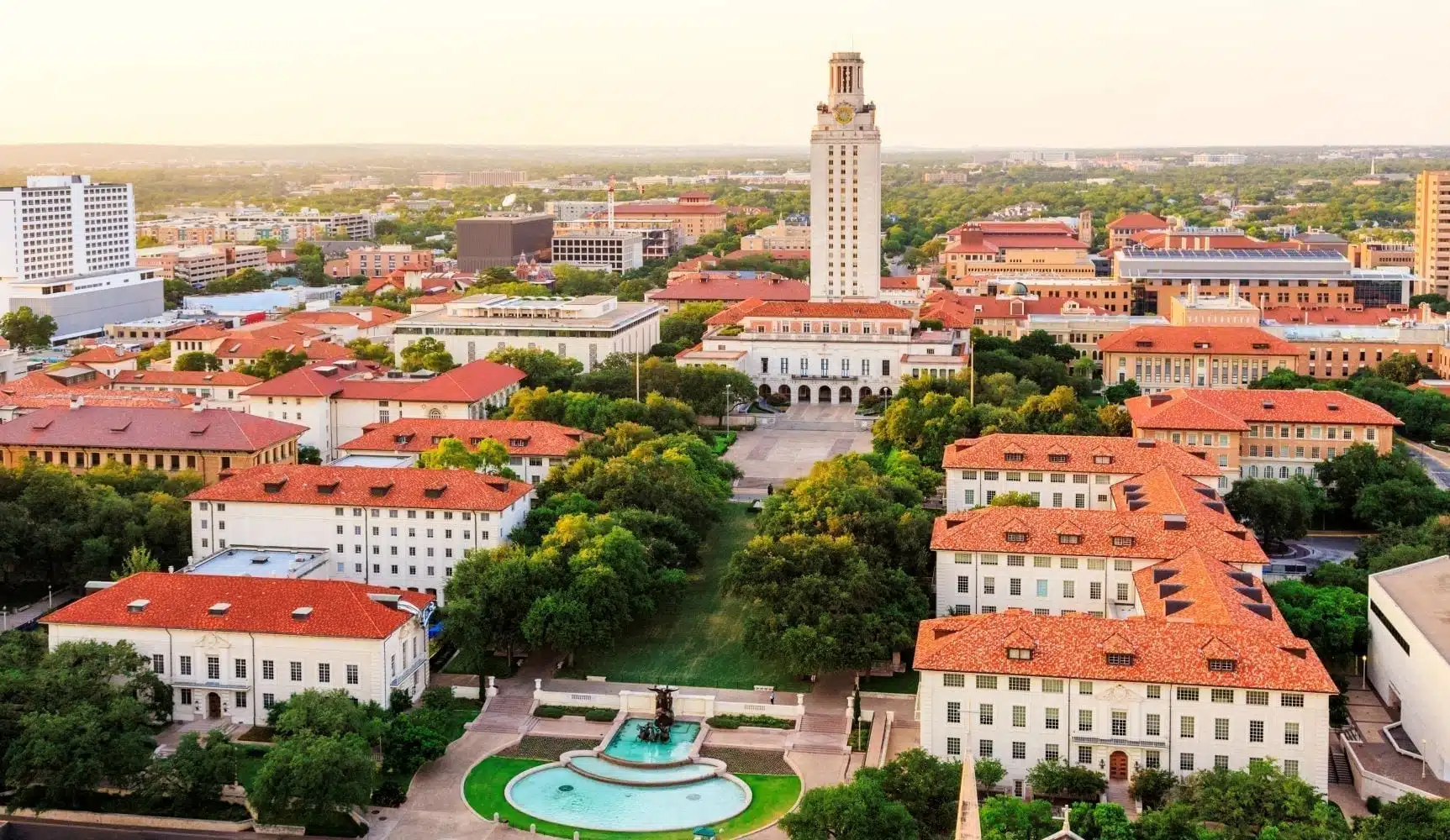 Aerial view of the University of Texas at Austin on a spring day. The large buildings on campus all have red tile roofs, and there is a tall tower rising up from one of the buildings. In the foreground is a three-tiered fountain and a green lawn, and there are mature trees casting shade between buildings.