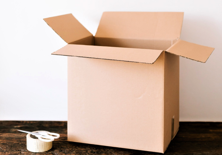 A cardboard box with its top open is sitting on a wooden table beside a roll of packing tape and a pair of scissors.