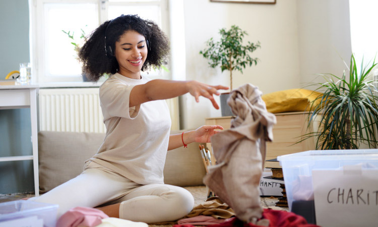 A young woman is listening to music on her headphones as she tosses a piece of clothing into a plastic bin labeled “Charity.” She’s decluttering her wardrobe and other belongings before she begins packing for college.