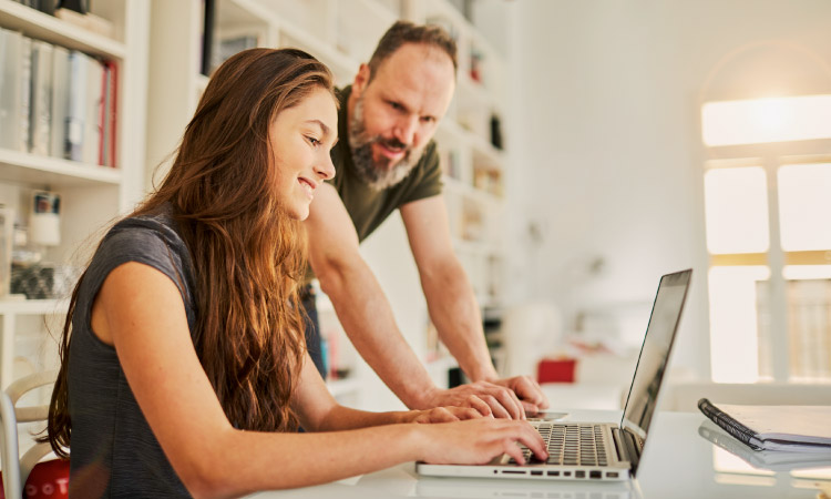 A high school senior is sitting in front of her laptop, making plans for her first year of college. Her father is standing nearby, helping her with planning, and they’re both smiling.