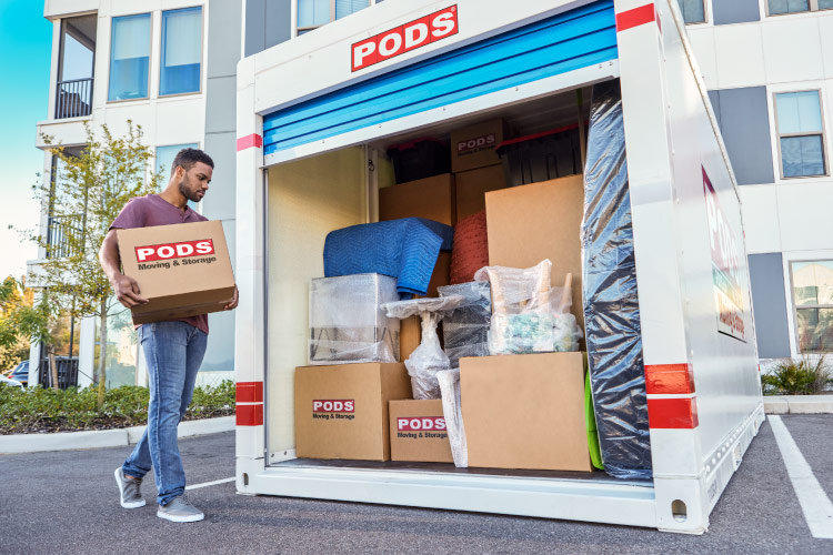 A young man is carrying a cardboard box (with the PODS Moving and Storage logo on it) to a PODS container positioned in the parking area of his apartment complex. The container’s blue door is open, and there are neatly stacked moving boxes, a mattress, and various pieces of wrapped furniture inside the container.