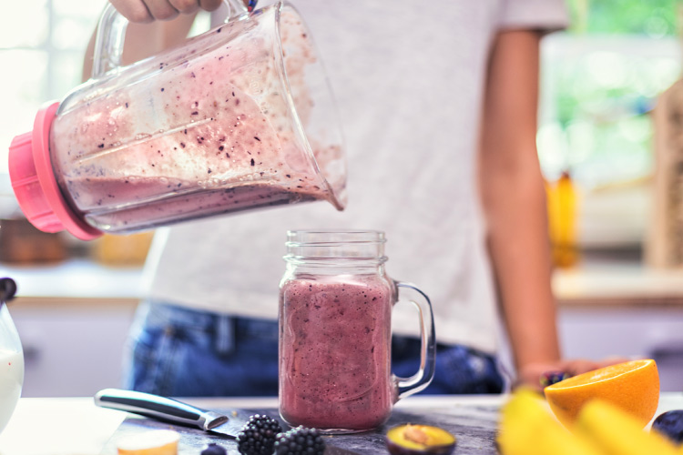 Close-up view of a young woman pouring a fruit smoothie into a glass drinking jar. She’s wearing jeans and a gray T-shirt. There are blackberries and various cut fruit on the counter, as well as the knife that was used to cut the fruit.