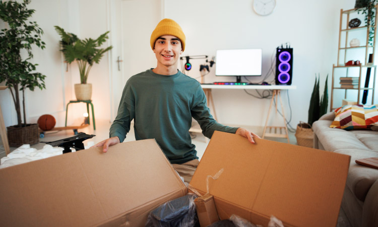 A young man in a yellow beanie is smiling excitedly at the camera as he holds open a large cardboard box with nondescript contents. The room behind him contains a pull-out couch, shelves, house plants, and a desk with a computer and light-up speakers.