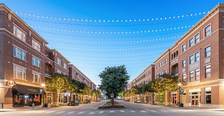 A four-lane road in Frisco Square with a tree-lined median. Apartments sit atop of ground-level stores, making it a great place to live, shop, and eat.