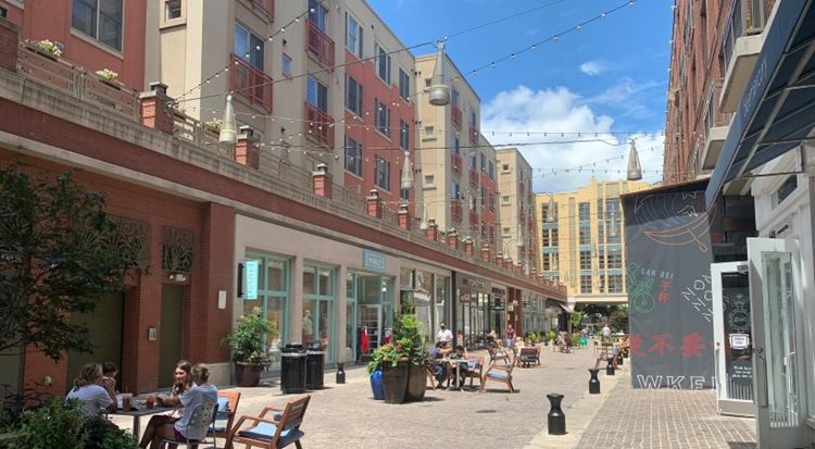 View of a downtown street in Bethesda, Maryland. Locals are out and about, enjoying a sunny day in the city. Some are dining al fresco, while others are strolling by storefronts.