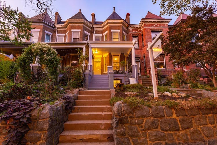 The view looking up at a majestic brownstone in the Mount Pleasant neighborhood of Washington, D.C. The home features two sets of steps leading up from the sidewalk, a stone wall beneath the lawn, and a classic Victorian look. 