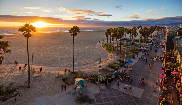 Aerial of Venice Beach, California, during sunset. Tourists and locals walk in the sand and along the boardwalk. 