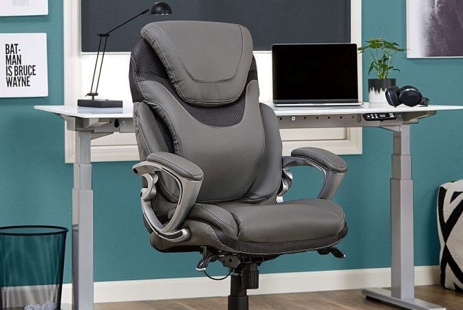 A high-end office chair from Serta. It has padding built into the chair to make it more comfortable. Behind the chair is a home office desk with a laptop, lamp, headphones, and a houseplant. 