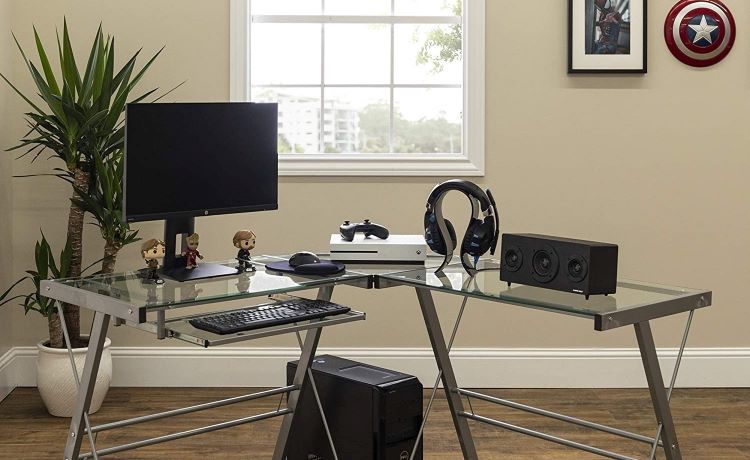 A glass and metal corner office desk used in a work-from-home office setup. There’s a large monitor, keyboard, speakers, headphones, a gaming station, and some bobbleheads on the desk. 