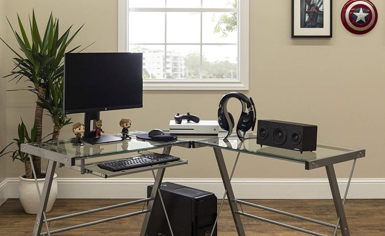 A glass and metal corner office desk used in a work-from-home office setup. There’s a large monitor, keyboard, speakers, headphones, a gaming station, and some bobbleheads on the desk.