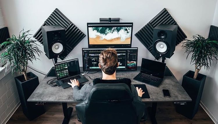 A man is working at his desk in his home office. He has dual monitors, two laptops, and high-end speakers with sound-dampening panels installed on the wall directly behind them.