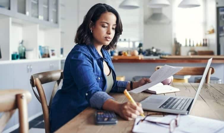 A woman is sitting in her kitchen, working out the budget for her home office. She’s writing notes on a piece of paper and has her laptop and a calculator nearby.