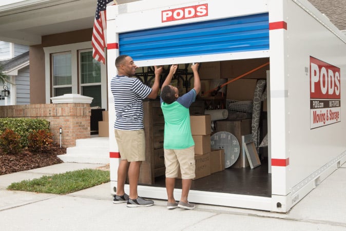 A man and his son opening the blue door on their PODS moving and storage container. It’s still half filled with boxes and various household items.