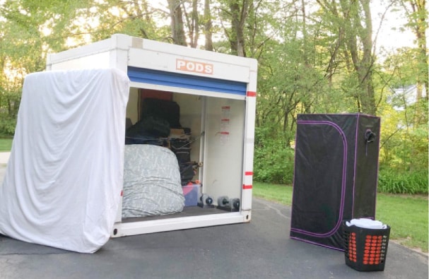 A PODS 8-ft portable moving container is sitting in a residential driveway with its door open. It’s nearly half filled with various household things. There’s a mattress leaning against the outside of the container and a couple of other items nearby, waiting to be loaded.