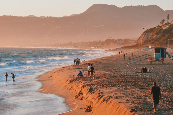 Locals enjoy a sunset at Point Dume in Malibu, California. The ocean is crashing against the sand, and there’s a small ledge where the dry beach drops down into the wet surf. There’s a lifeguard stand on the beach and cliffside houses in the background.