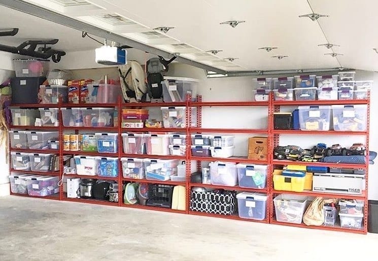 Red metal shelves line a garage wall. It is filled with different bins filled with items from the garage. 