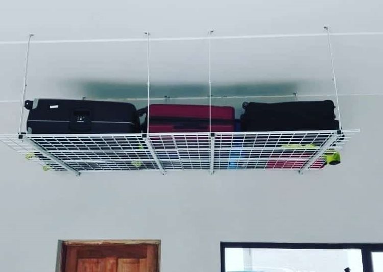 A white storage rack hangs from the ceiling of a garage. It is filled with suitcases, two black and one pink. 