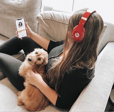 A woman sitting on her couch with a dog on her lap while listening to an audiobook through red headphones