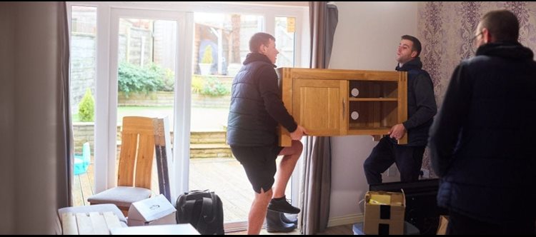 Two men are carrying part of a media console while another man is standing nearby, waiting to pick up the next piece of furniture and take it to the moving truck.