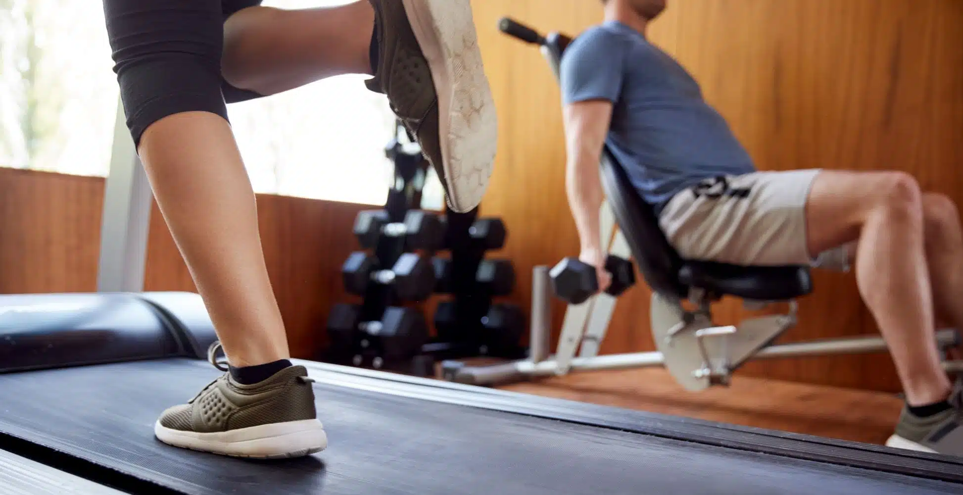 Floor-level view of a home gym with two people working out. A man is sitting on a bench, lifting dumbbells, and a woman is running on a treadmill in the foreground.