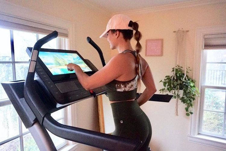 A young woman in workout attire is using a high-tech treadmill with a touch screen. The treadmill is set up in her home gym.