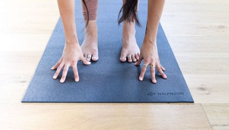 Close-up view of a woman’s feet and hands on a yoga mat as she does a forward fold.