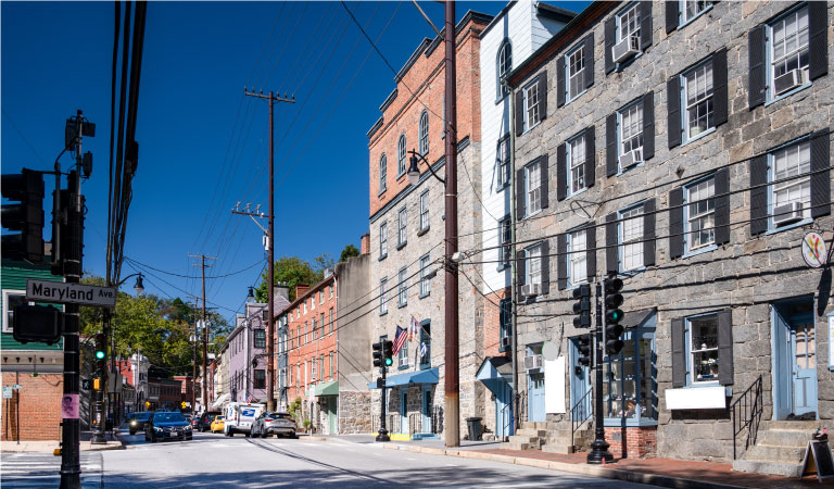 Street-level view of the shopping district in historic Ellicott City, Maryland. A row of old, brick buildings lines the street. There are a few cars parked along the curb and others are about to cross an intersection. A street sign on the left side of the image reads, “Maryland Ave.”