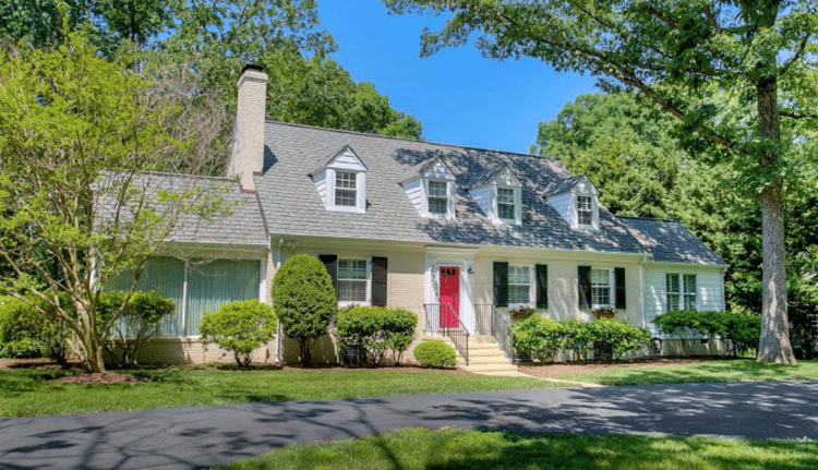 A lovely, two-story home in the Stratford Hills neighborhood of Richmond, Virginia. The home is painted a light beige color with black shutters and a bright red door. There are several mature trees surrounding the house, and some nicely shaped bushes line the front side.