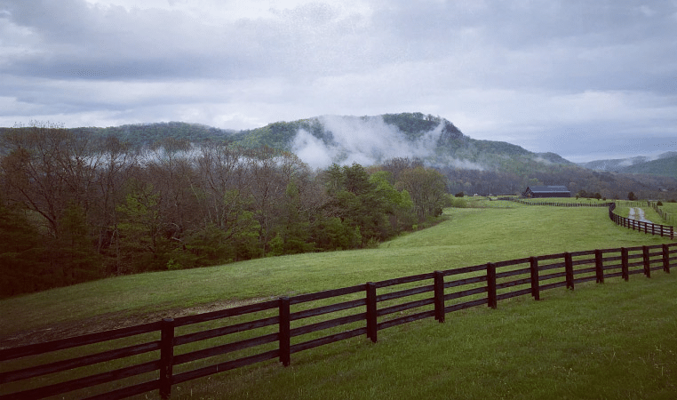 The forests and mountains around Berea, Kentucky, in the early morning. A misty fog is rolling in under a cloudy sky. In the foreground are mowed fields and a long, wooden fence, and in the distance is a large barn.