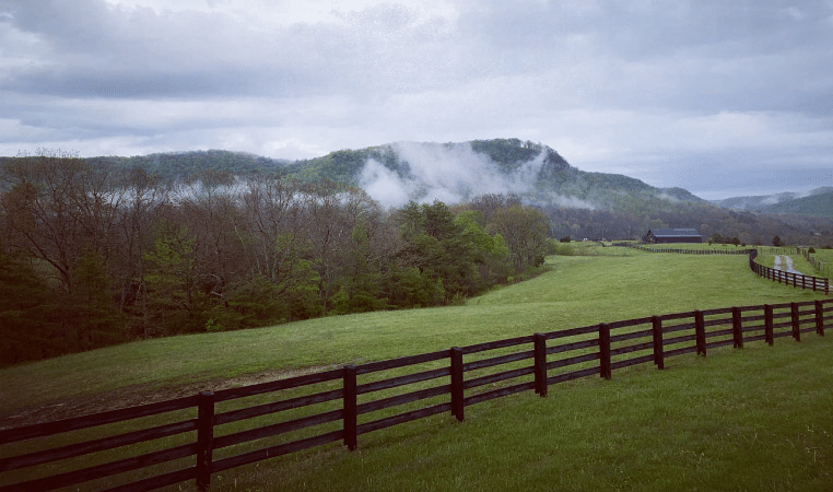 The forests and mountains around Berea, Kentucky, in the early morning. A misty fog is rolling in under a cloudy sky. In the foreground are mowed fields and a long, wooden fence, and in the distance is a large barn. 