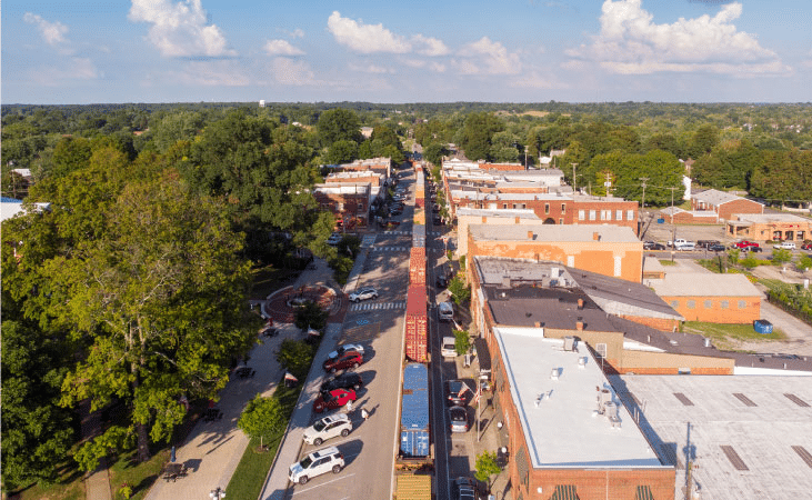 Aerial view of the town of La Grange in Kentucky as a freight train travels through the downtown area. Surrounding the town are mature trees filled to the brim with lush, green leaves. 