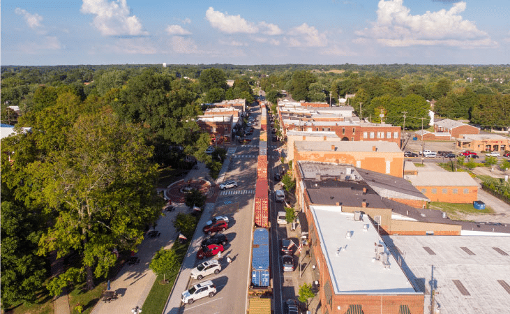 Aerial view of the town of La Grange in Kentucky as a freight train travels through the downtown area. Surrounding the town are mature trees filled to the brim with lush, green leaves.