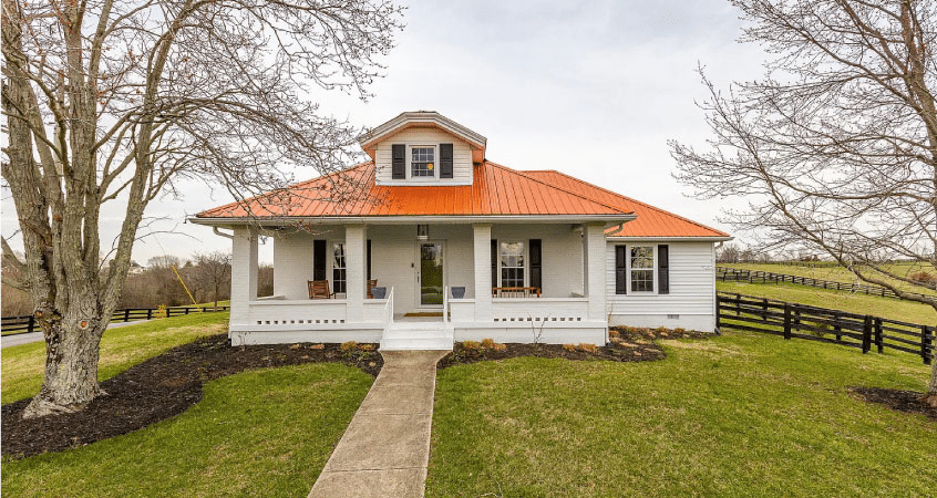 A beautiful one and a half story country house in Harrodsburg, Kentucky. It has a large covered porch and an orange metal roof. The home is white with black shutters and is surrounded by an expansive lawn and a couple mature trees. 
