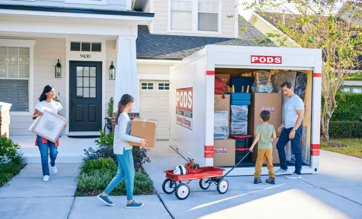 A family of four is carrying things out of their house and loading them into a PODS container in their driveway.