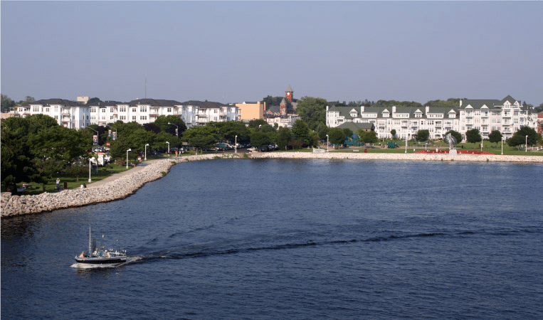 View of the harbor in Ludington, Michigan. There are large, white residential buildings close to the shore and a sailboat is motoring away from the town. 