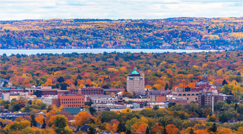 Distant view of Traverse City, Michigan, in the fall. The trees are a plethora of hues from dark green to yellow, orange, and red. Downtown Traverse City stands out among the vast foliage and water cuts across the landscape in the distance.