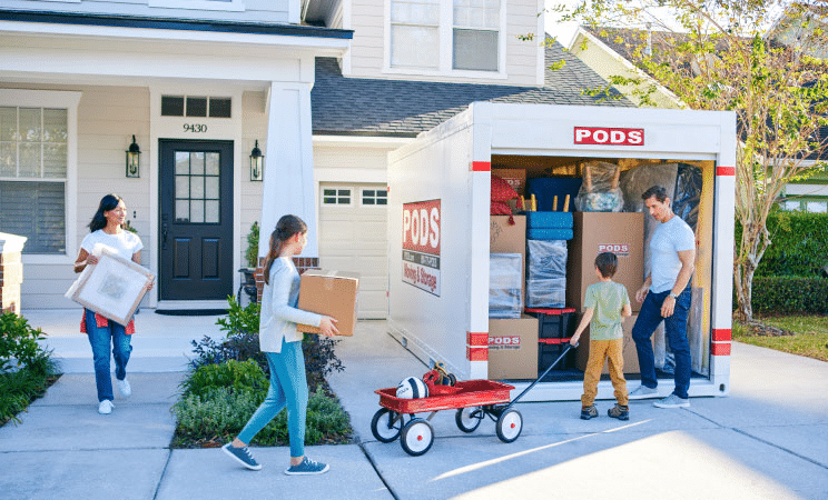 A family of four is working together to load the last of their things into a PODS portable moving container in their driveway. The father is standing by the container speaking with his son who is pulling a red wagon. The daughter is carrying a moving box and the mother is carrying a framed picture.