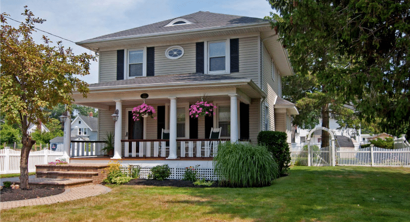 A two-story residential home in Islip, New York, on Long Island. The home is made of beige siding and there are black shutters on either side of each window. There’s a large covered porch with doric columns holding up the first floor roof. 