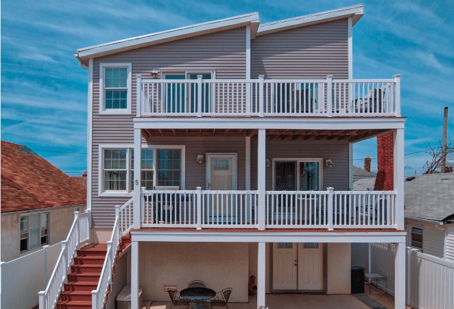 A three-story beach house in Long Beach, New York, on Long Island. The second and third stories have wraparound decks with the lower one extending beyond the bottom floor to create a covered deck. Red stairs with white railings lead up from the ground floor to the second level.