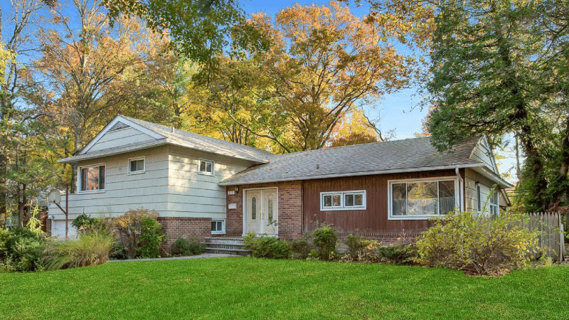 A brick and siding single-family home in Great Neck, New York. The second level is below ground and the home sits in the middle of several mature trees and large, green lawn. 