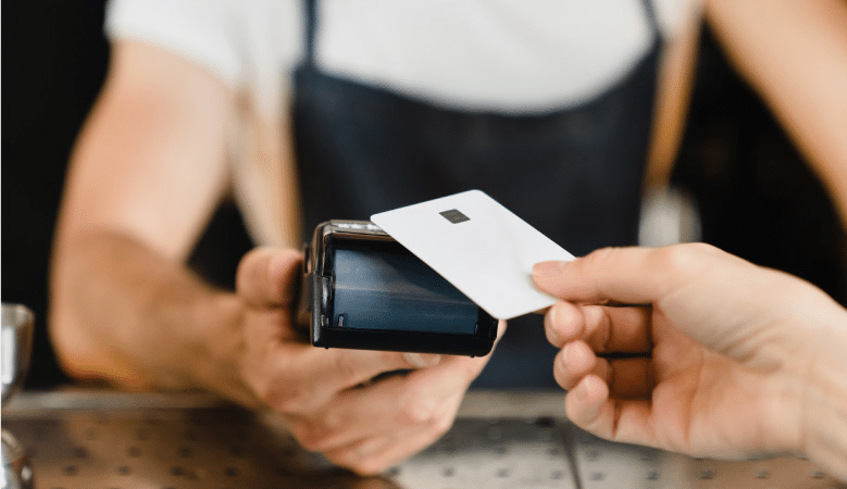 Close-up shot of a woman wearing a black apron and a white shirt and holding a wireless card reader as a customer taps their credit card to make a payment. 